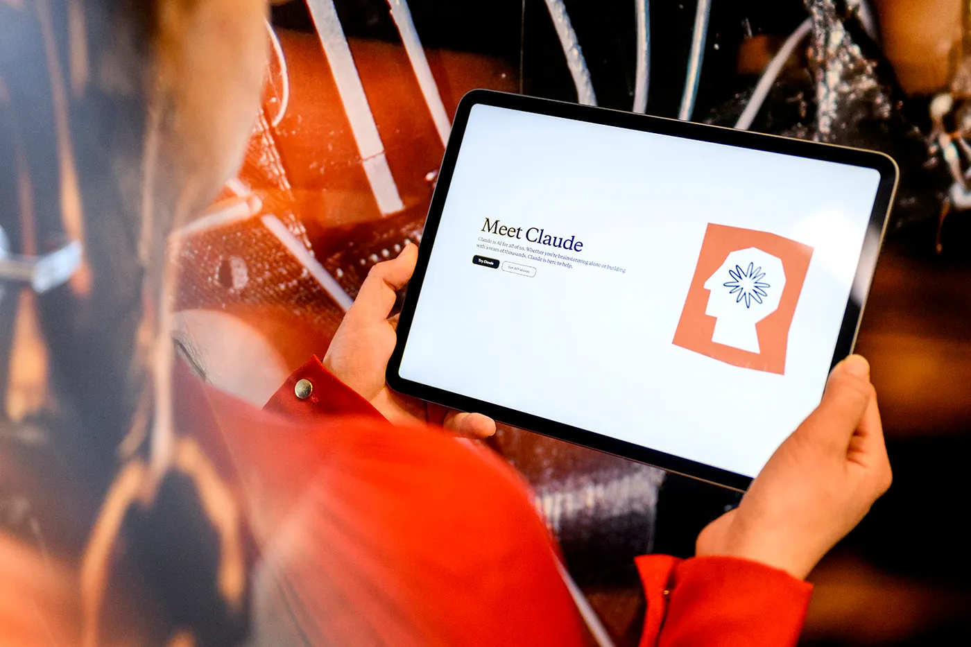 A person in a red shirt holds a tablet displaying a webpage titled "Meet Claude," featuring a logo of a head profile with a starburst symbol. The background contains out-of-focus, warm-toned lights and patterns.