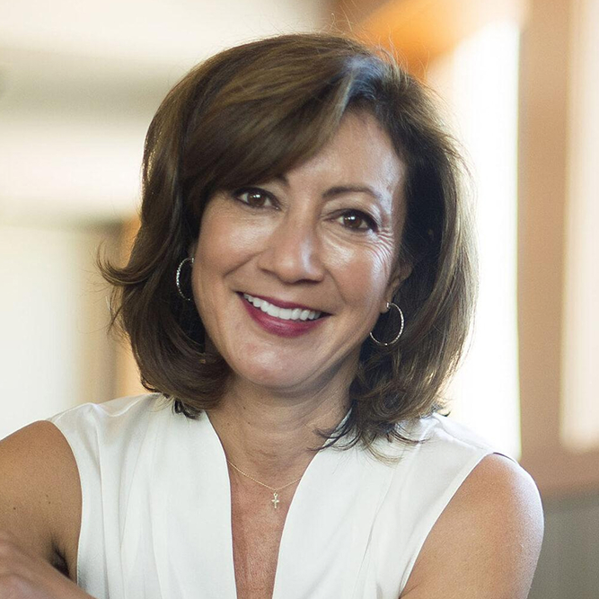A woman with shoulder-length brown hair, wearing a sleeveless white top and hoop earrings, smiles warmly at the camera in a softly lit indoor setting.
