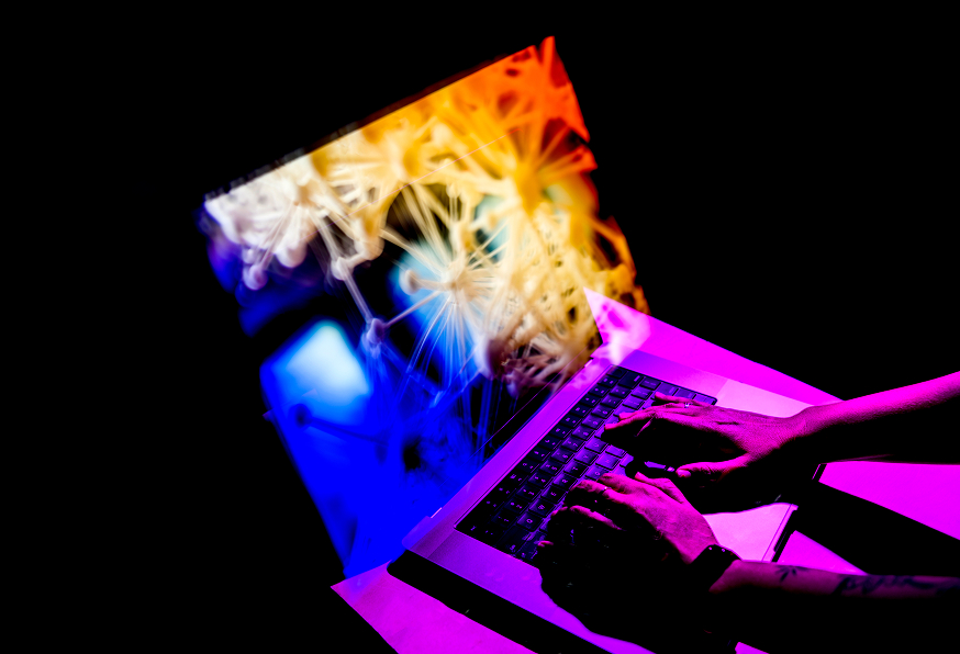 Hands typing on a laptop keyboard with a colorful, abstract neural network pattern projected from the screen, creating a vivid blend of blue, yellow, and pink light in a dark setting.