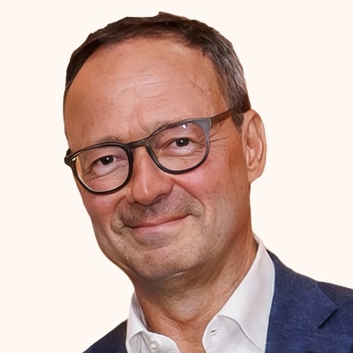 A middle-aged man with short brown hair, wearing round eyeglasses, a white shirt, and a blue blazer, smiling at the camera against a light background.