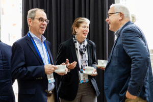 Three people in business attire smiling and talking while holding coffee cups at a professional event, standing in front of dark curtains.