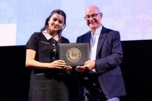 A woman and a man stand side by side on a stage, smiling and holding a certificate with a university seal. The background features a blue and white floral pattern.